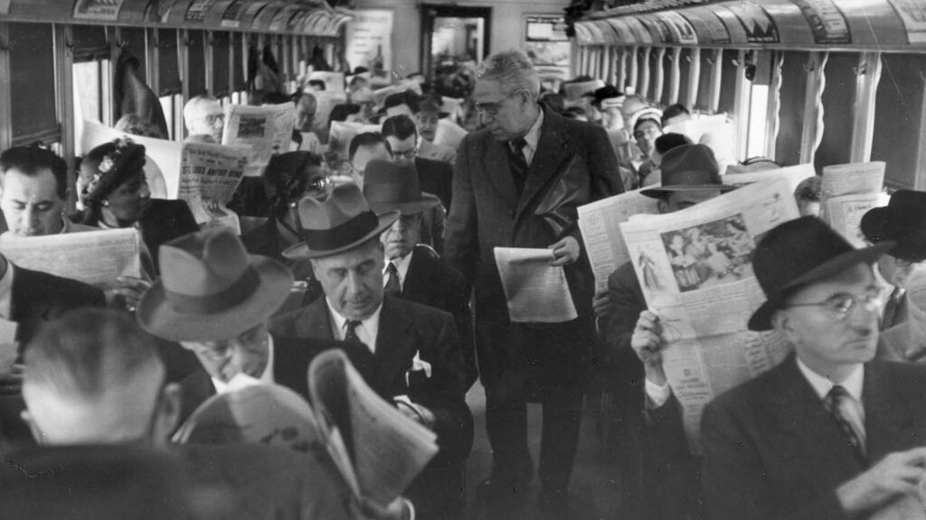 Commuters reading broadsheet newspapers on a packed train
